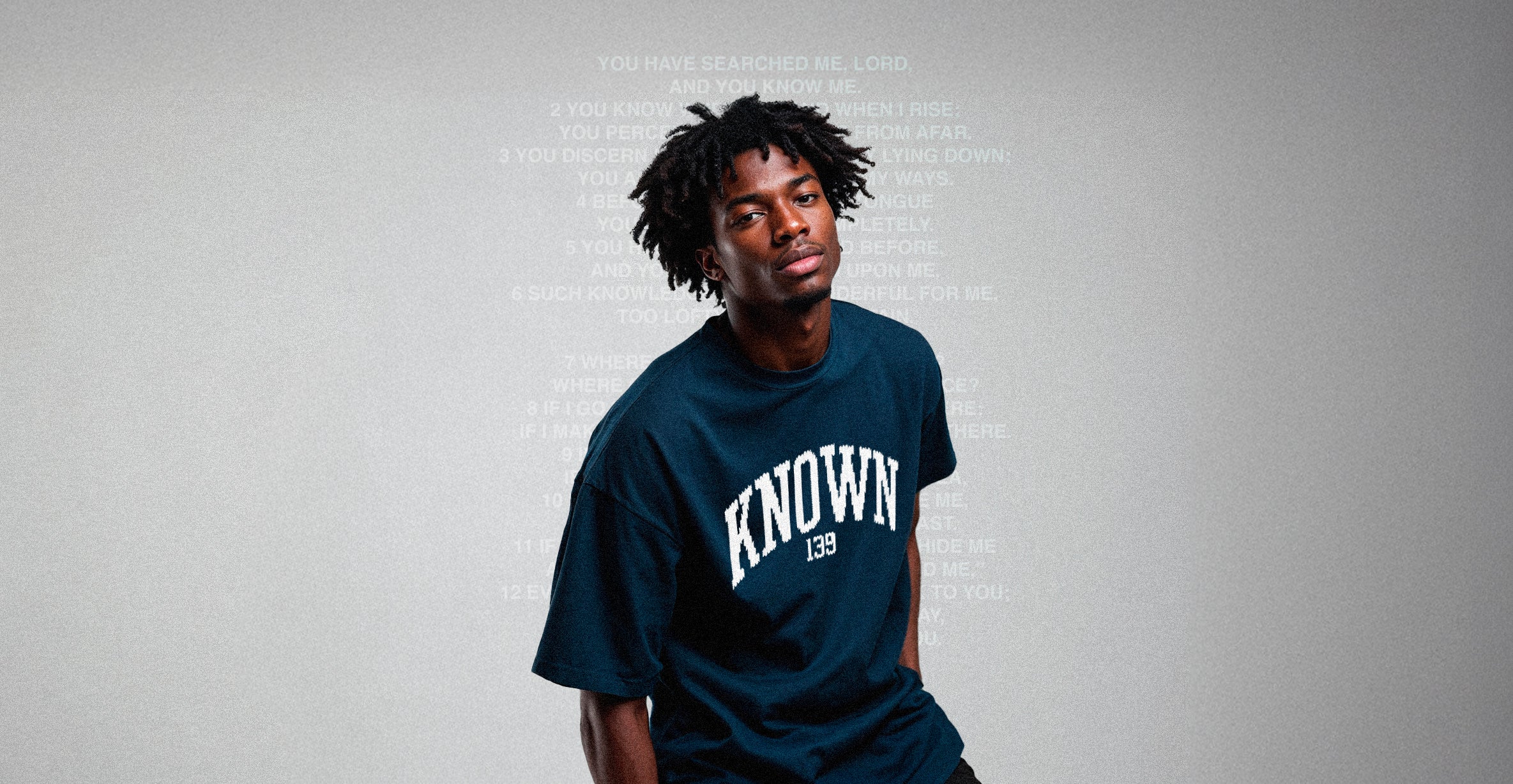 Christian apparel UK streetwear. Person wearing a navy blue t-shirt with 'KNOWN' printed on it against a gray background.